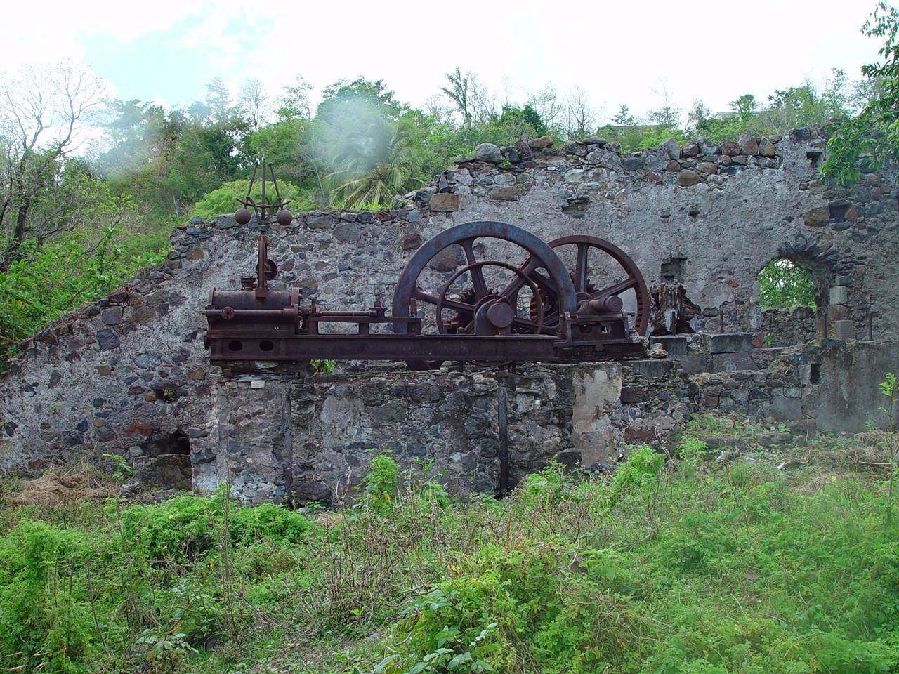 Vestiges de la machine à vapeur de Clairefontaine : deux grands volants de fonte rouillée et leur bâti, restés en place au sommet d'un mur de pierres, au milieu des ruines à ciel ouvert envahies par la végétation.
