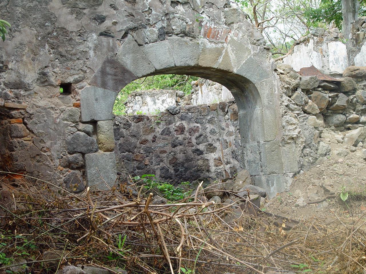Porte en plein cintre appareillée en pierre de taille, ouverte dans un mur de moellons volcaniques en ruine, donnant sur la végétation.
