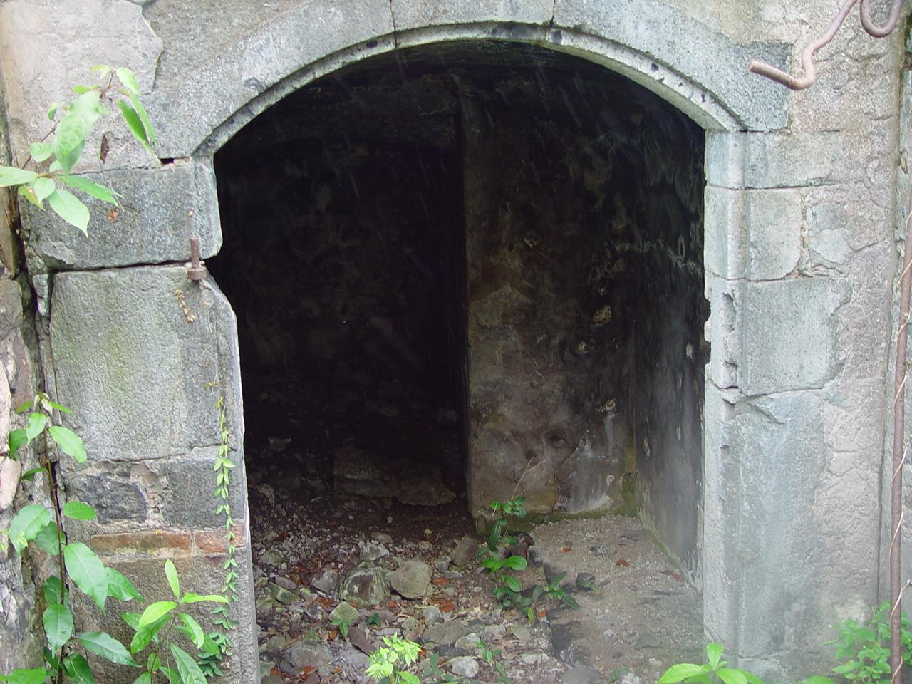 Ouverture voûtée en pierre de taille donnant sur une salle basse et obscure, au sol jonché de pierres ; des gonds de fer rouillés sont encore scellés dans les piédroits.